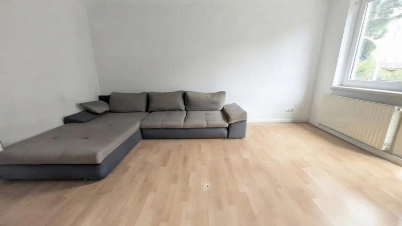Helles Wohnzimmer mit Ecksofa, Parkettboden und großem Fenster, das viel Tageslicht hereinlässt.