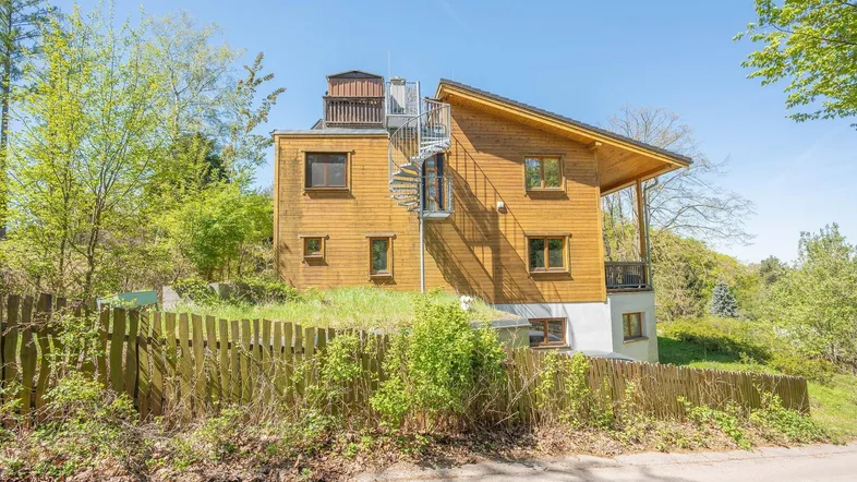 Holzhaus mit Wendeltreppe an der Fassade und umgebender Natur unter blauem Himmel.