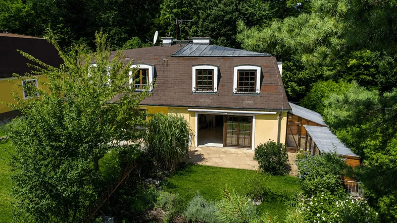 Gelbes Haus mit braunem Mansarddach, umgeben von üppigem Grün und einem kleinen Gartenbereich.