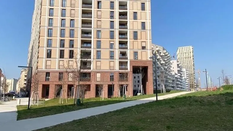 Hohes modernes Wohngebäude mit vielen Balkonen und einer markanten Ziegelfassade im unteren Bereich unter blauem Himmel.