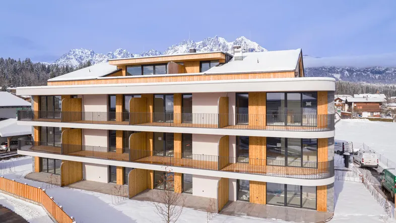 Modernes Apartmentgebäude mit Holzfassade und Balkonen vor verschneiter Bergkulisse.