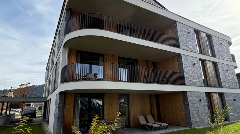 Modernes Mehrfamilienhaus mit geschwungenen Balkonen und Steinverkleidung in alpiner Umgebung.