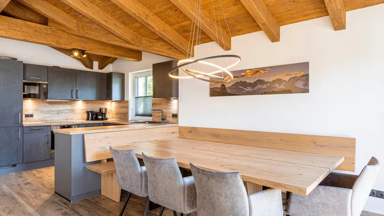 Moderne offene Küche mit dunklen Schränken, Holzelementen und einem großen Esstisch unter Holzbalkendecke.