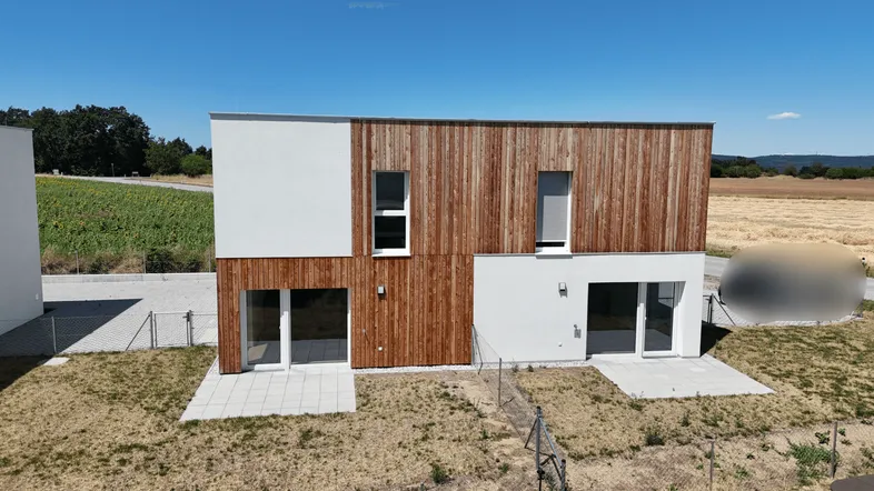 Moderne Doppelhaushälfte mit Holz- und Putzfassade, umgeben von Feldern unter blauem Himmel.
