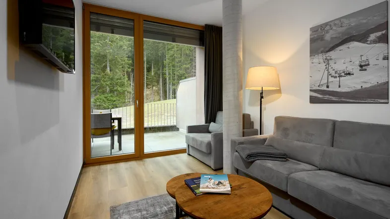 Helles Wohnzimmer mit gemütlichem Sofa, Holzboden und Zugang zu einem Balkon mit Waldblick.