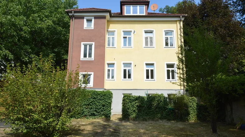 Gelbes Mehrfamilienhaus mit braunem Anbau und grüner Bepflanzung im Vorgarten.