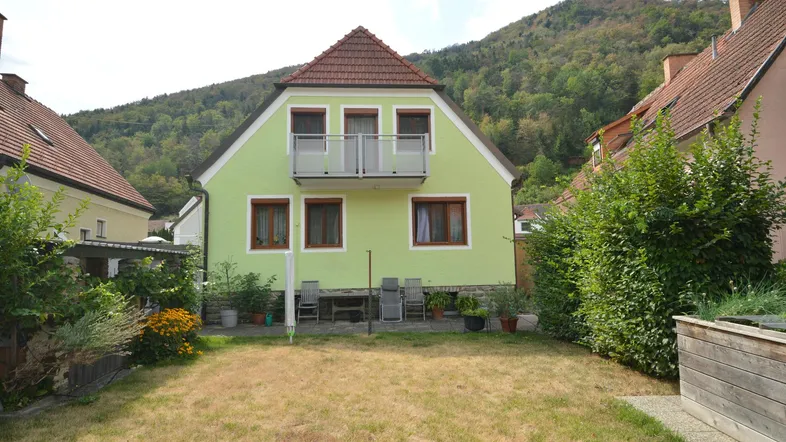Hinteransicht des Hauses mit hellgrüner Fassade, kleinem Balkon und Gartenbereich vor bergiger Kulisse.