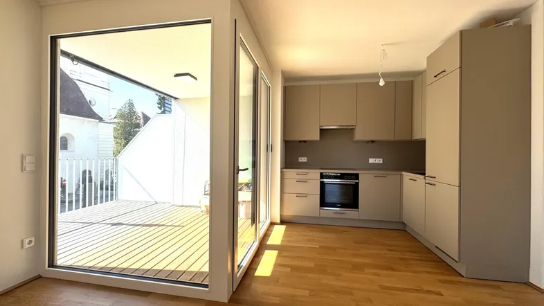 Heller offener Wohnbereich mit moderner Einbauküche, Holzboden und Zugang zu einer sonnigen Terrasse.