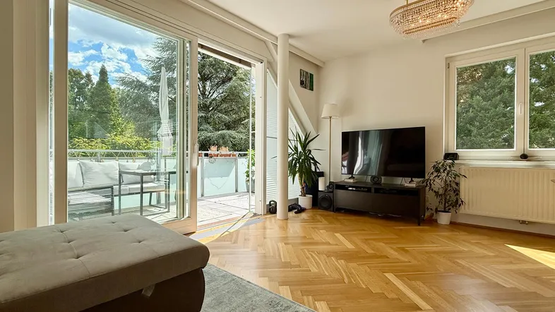 Helles Wohnzimmer mit Parkettboden, Zugang zum Balkon und weitem Blick ins Grüne.