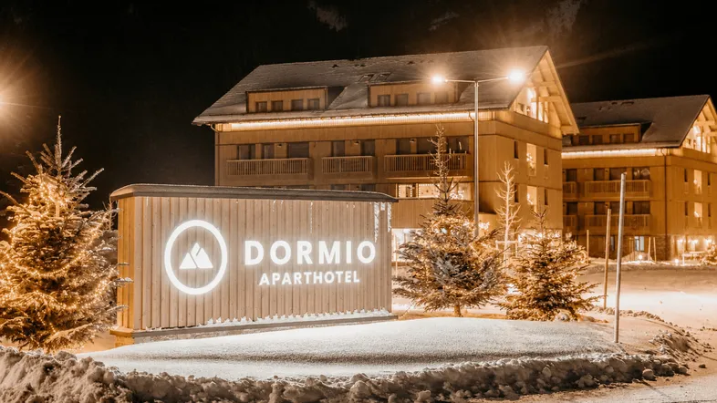 Beleuchtetes DORMIO Aparthotel Schild im Schnee bei Nacht, umgeben von festlich geschmückten Tannenbäumen.