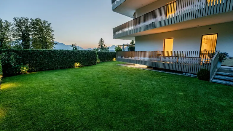 Gepflegter Gartenbereich mit Rasenfläche, Hecke und Blick auf die Berge bei Sonnenuntergang.