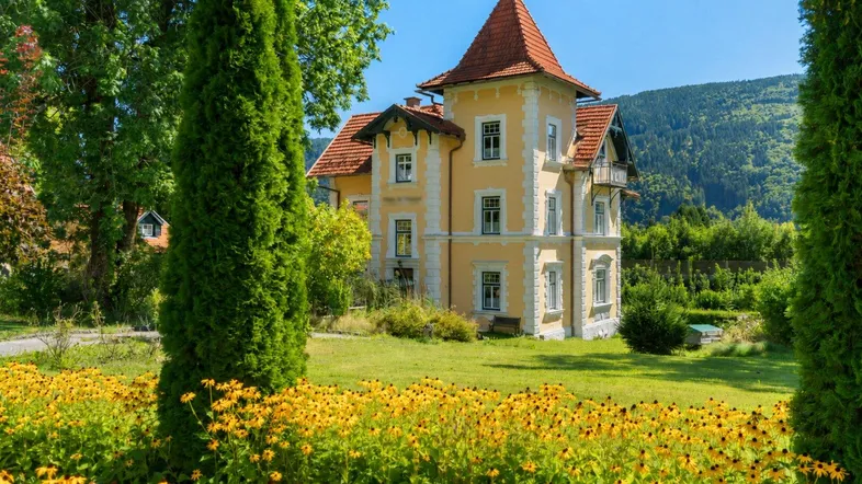 Historische Villa mit markantem Turm und weitläufigem Garten, umgeben von blühenden Blumenbeeten und hohen Bäumen.