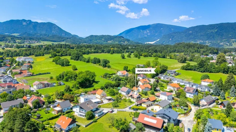 Luftaufnahme einer Wohnsiedlung mit grünen Feldern und Bergen im Hintergrund, die Immobilie ist markiert.