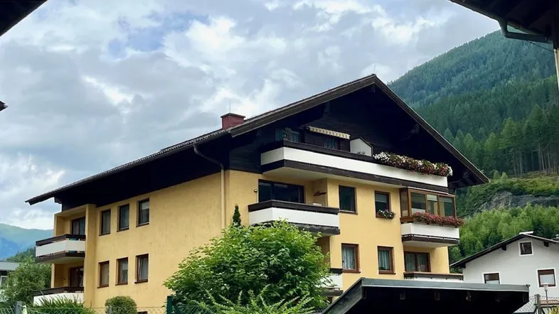 Mehrfamilienhaus in alpiner Umgebung mit Balkonen und Satteldach, umgeben von grünen Bäumen.
