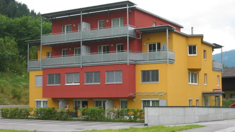 Modernes Mehrfamilienhaus mit leuchtend rot-gelber Fassade und mehreren Balkonen in grüner Umgebung.
