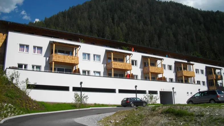 Modernes Mehrfamilienhaus mit Balkonen und Parkplätzen vor einer bewaldeten Bergkulisse.