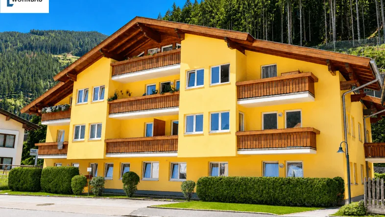 Gelbes Mehrfamilienhaus mit Holzbalkonen und Satteldach vor bewaldeter Bergkulisse.
