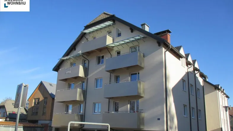 Modernes Mehrfamilienhaus mit heller Fassade, mehreren Balkonen und Satteldach unter blauem Himmel.