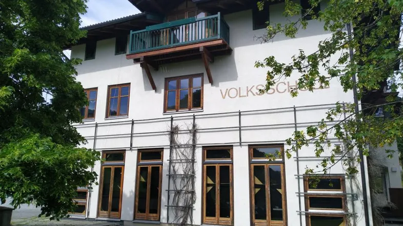 Historisches Gebäude mit weißer Fassade, Holzbalkon und grüner Umgebung, ehemals eine Volksschule.