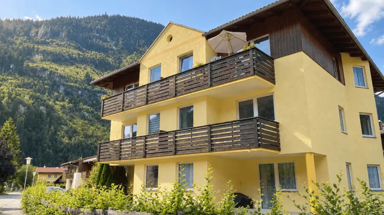 Gelbes Mehrfamilienhaus mit Holzbalkonen und Bergblick in sonniger Umgebung.