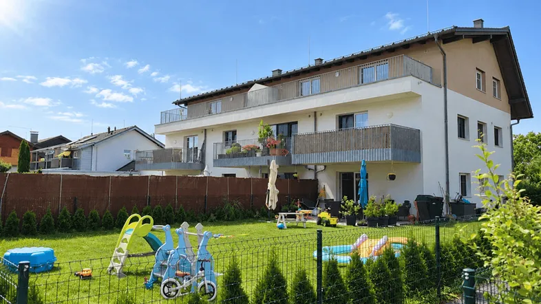 Gepflegter Gartenbereich mit Spielplatz und einem modernen Mehrfamilienhaus im Hintergrund unter blauem Himmel.