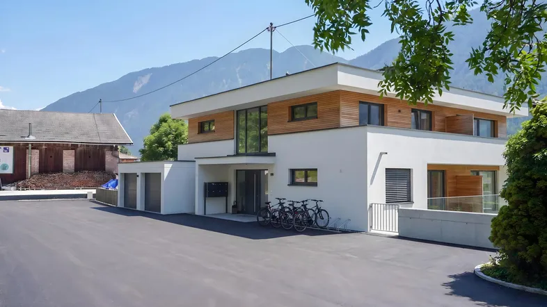 Moderne Architektur mit weißer Fassade, Holzverkleidung und großen Fenstern vor alpiner Kulisse.