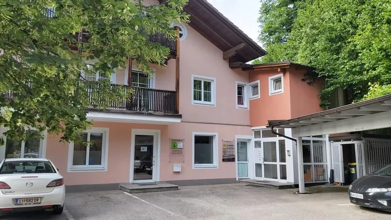 Ansicht des Gebäudes mit mehreren Eingängen, Parkplätzen und umgebender Vegetation.