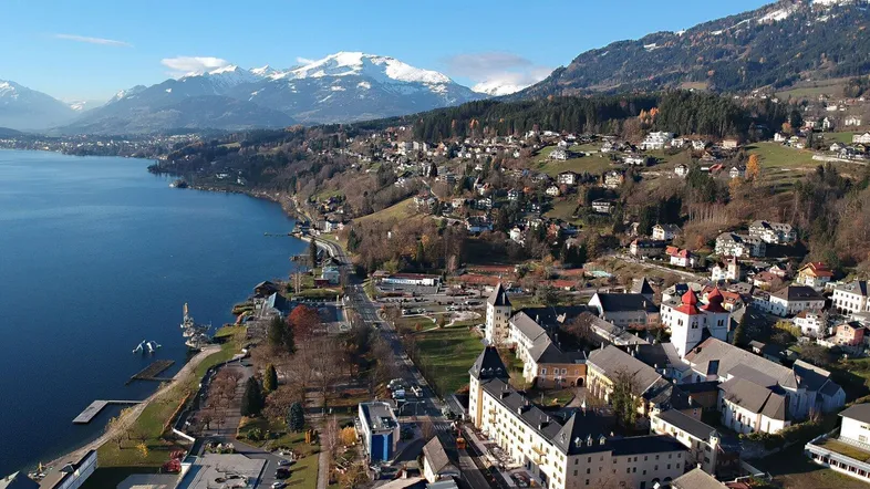 Panoramablick auf eine malerische Stadt am Seeufer mit schneebedeckten Bergen im Hintergrund unter blauem Himmel.