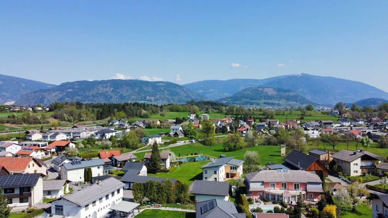 Panoramablick über eine Wohnsiedlung mit grünen Feldern und Bergen im Hintergrund unter blauem Himmel.