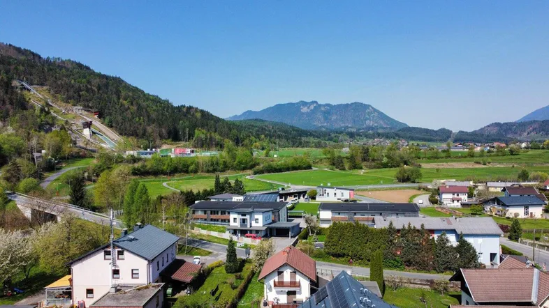 Luftaufnahme einer Wohnsiedlung mit grüner Landschaft und Bergen im Hintergrund unter blauem Himmel.