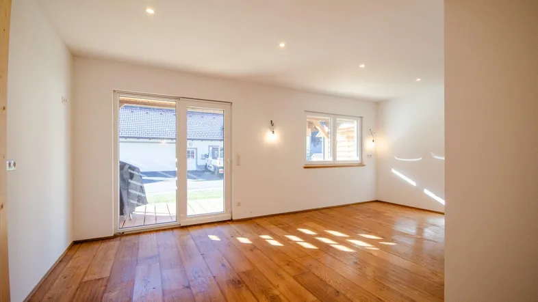 Helles, leeres Zimmer mit Holzboden, großen Fenstern und Zugang zur Terrasse.