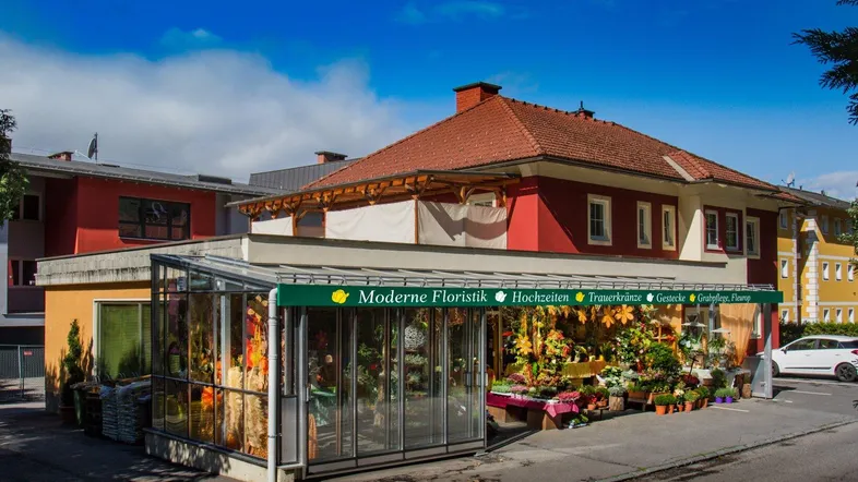 Außenansicht eines Blumengeschäfts mit großem Glasanbau und rotem Dach unter blauem Himmel.