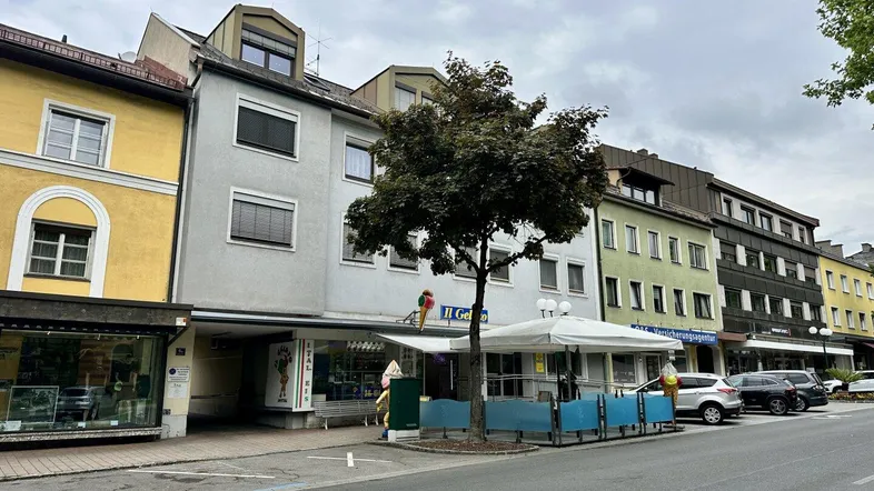 Ansicht einer belebten Straße mit mehreren Gebäuden, Geschäften im Erdgeschoss und parkenden Autos.