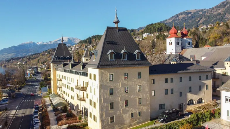 Historisches Gebäude mit Türmen und Blick auf Berge und See, umgeben von einer Straße und Bäumen.