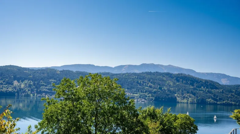 Panoramablick auf den See und die umliegenden bewaldeten Berge unter strahlend blauem Himmel.