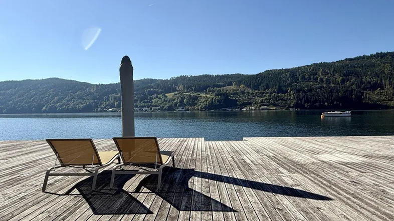 Große Holzterrasse am See mit Liegestühlen und Bergblick unter blauem Himmel.
