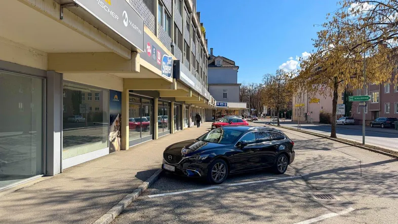Gebäudeansicht mit Geschäften im Erdgeschoss und parkenden Autos auf der Straße unter blauem Himmel.