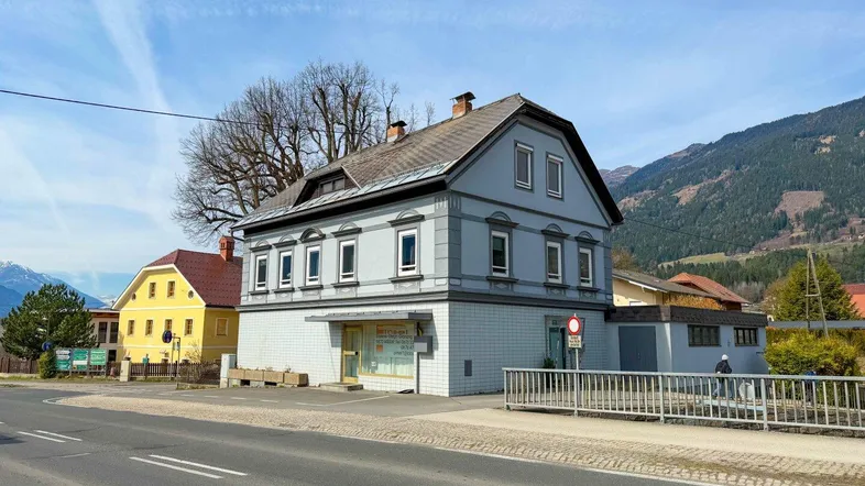 Historisches Gebäude mit grauer Fassade und Mansarddach an einer Straße, umgeben von Bergen.