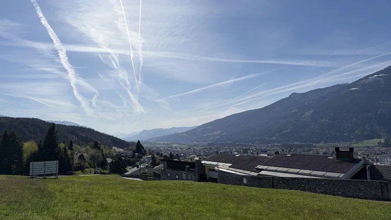 Blick über eine grüne Wiese auf eine Siedlung mit Bergen im Hintergrund unter blauem Himmel.