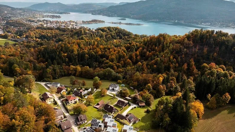 Panoramablick auf eine Siedlung am Seeufer mit herbstlichen Wäldern und Bergen im Hintergrund.