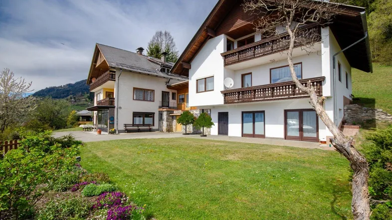 Traditionelles Haus mit gepflegtem Garten und Balkonen, umgeben von grüner Landschaft und Bergen.