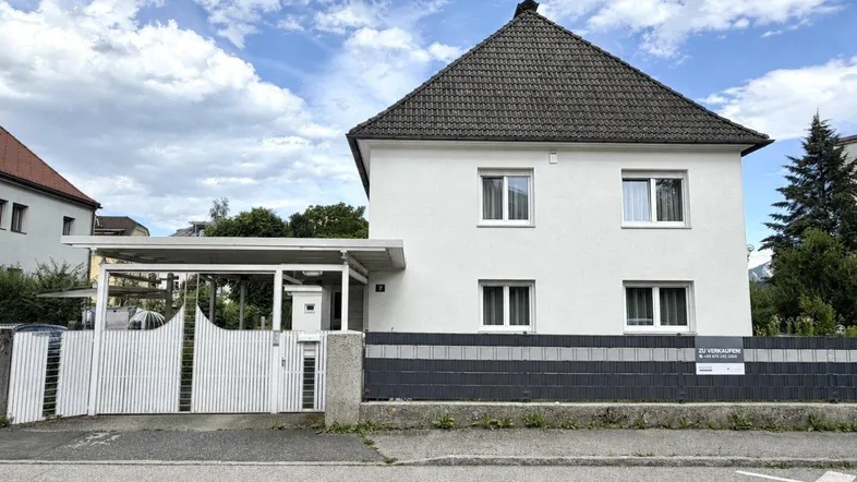 Weißes Einfamilienhaus mit Satteldach, Carport und Zaun, bereit zum Verkauf.