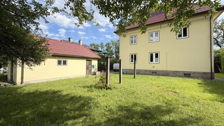 Großer Gartenbereich mit Rasenfläche und einem Nebengebäude, umgeben von Bäumen unter blauem Himmel.