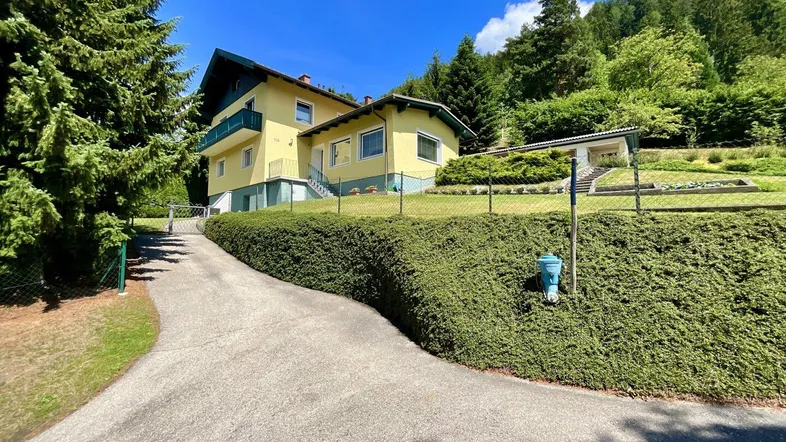 Gelbes Wohnhaus mit grüner Hecke und Auffahrt, eingebettet in eine hügelige Landschaft mit Bäumen.