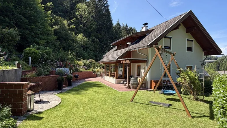 Gepflegter Gartenbereich mit Rasenfläche, Schaukel und Grillplatz vor dem Haus mit Waldblick.