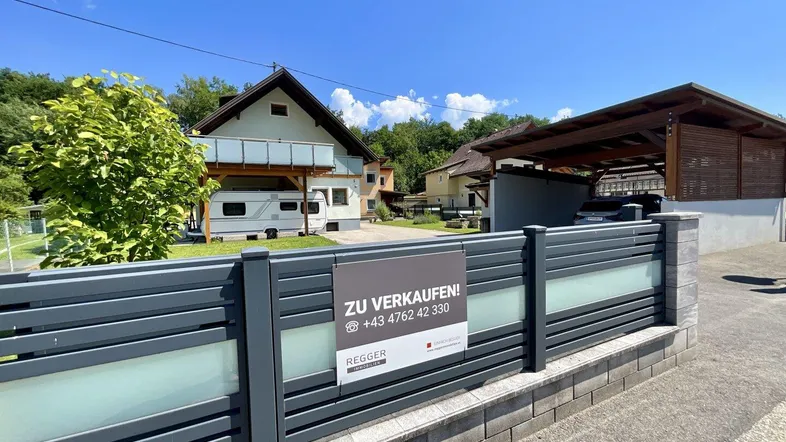 Außenansicht des Hauses mit modernem Zaun, Carport und einem Verkaufsschild im Vordergrund.