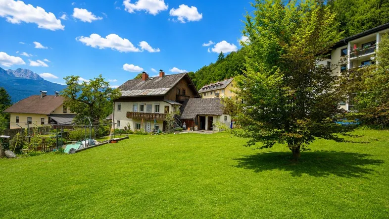 Weitläufige Grünfläche mit Blick auf das charmante Wohnhaus und die umliegende Berglandschaft unter blauem Himmel.