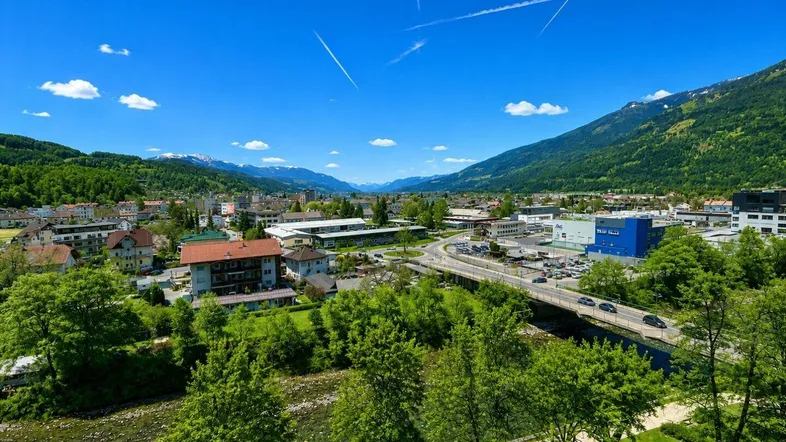 Weitläufiger Blick über eine Stadt mit Fluss, grünen Bäumen und schneebedeckten Bergen im Hintergrund.