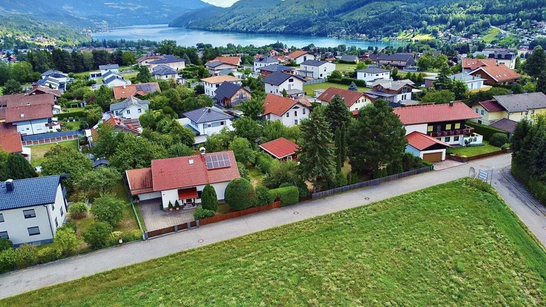 Luftaufnahme einer Wohnsiedlung mit Häusern, grünen Feldern, einem See und Bergen im Hintergrund.