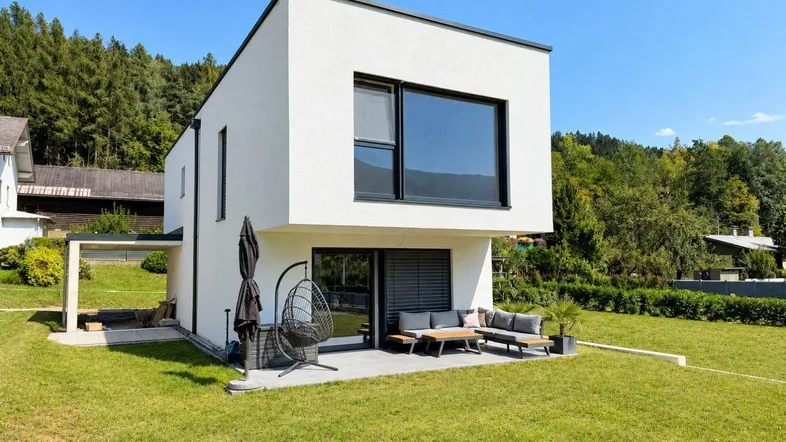Moderne weiße Villa mit Flachdach, großer Terrasse und Garten in grüner Umgebung.